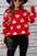 Red sweater with white heart pattern, long sleeves, round neck, paired with black jeans, modeled by a woman wearing a hat and sunglasses.